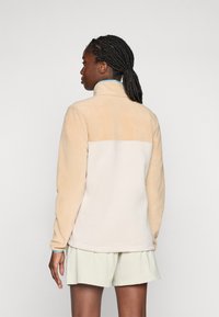 Vrouw met gevlochten haar, gekleed in een beige en crèmekleurige fleece jas en lichte shorts, staande met de rug naar de camera tegen een effen achtergrond.