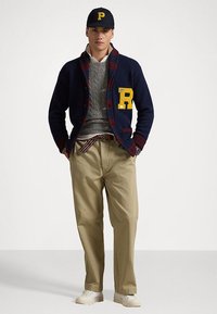 Cardigan en tricot marine avec des accents bordeaux, patch logo jaune ; pull en tricot gris en dessous ; pantalon kaki ; baskets blanches ; casquette noire.