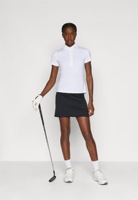 Camiseta polo branca com mangas curtas e abertura de três botões, combinada com uma saia-short preta. O modelo segura um taco de golfe, vestindo ténis brancos.