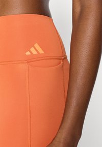 Πορτοκαλί κολάν με ψηλή μέση από ελαστικό ύφασμα, με πλαϊνή τσέπη και διακριτικό λογότυπο σε τονικό χρώμα στη πίσω μέση.
