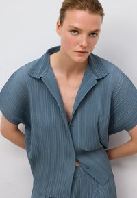 Camicia azzurra chiara, con maniche corte a pieghe, colletto con intaglio, chiusura a bottoni e vestibilità rilassata, caratterizzata da una texture a righe verticali e dettagli drappeggiati.