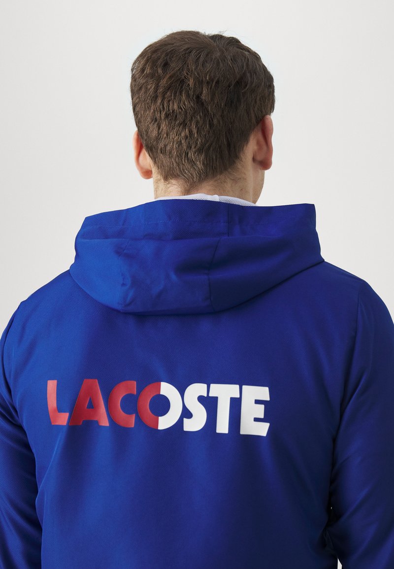 Giacca con cappuccio blu dal tessuto liscia, con un grande logo "LACOSTE" sulla schiena in rosso e bianco.