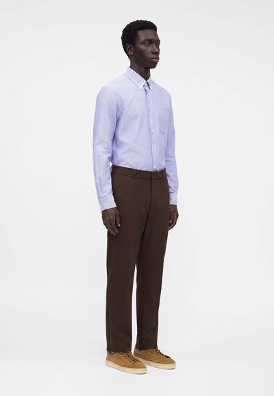 Un homme debout porte une chemise bleu clair boutonnée, un pantalon marron foncé et des baskets en daim beige sur un fond blanc uni.