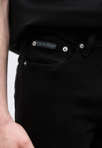 Κοντινό πλάνο στην τσέπη μαύρων τζιν με ετικέτα Calvin Klein και τέσσερις ασημένιους κρίκους, συνδυασμένα με μαύρο πουκάμισο.