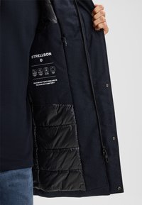 Giacca esterna blu navy con texture liscia, dotata di un interno trapuntato nero. Include etichette riflettenti per performance e comfort.