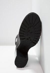 Czarna podeszwa buta o teksturowanej powierzchni, z głębokimi rowkami zapewniającymi przyczepność i lekko podwyższonym obcasem. Materiał wygląda na gumowany i trwały.