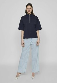 Sudadera navy con cuello alto y cremallera, mangas cortas y amplias, combinada con pantalones vaqueros de pierna ancha de color azul claro y tacones blancos de punta.