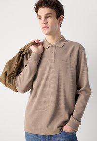 Beige langermet polo skjorte med ribbestruktur og krage, med tre knapper og en diskret "BOSS"-logo. Modell som holder en brun veske.