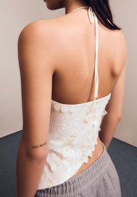 Žena v bílém, flitrovém, odkrytém topu na krk s tetováním na paži, v šedých kalhotách, zobrazena zezadu.