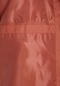 Le tissu de la veste couleur cuivre présente une texture lisse et brillante avec une couture horizontale et deux poches bordées dans le même matériau.