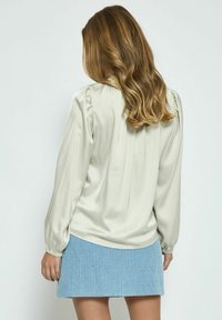 Blusa di raso beige chiaro con maniche lunghe, caratterizzata da pieghe sul retro, abbinata a una gonna plissettata blu chiaro con texture.
