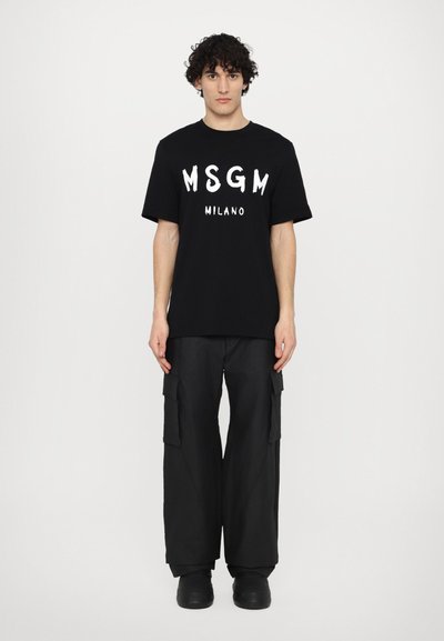 Musta puuvillapaita, jossa on valkoinen "MSGM MILANO" -painatus, yhdistettynä löysiin mustiin cargo-housuihin ja mustiin liukumasuksiin.