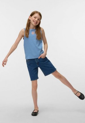 Smiling girl in blue striped sleeveless top and denim shorts balancing on one leg wearing black ballet flats on white background.
