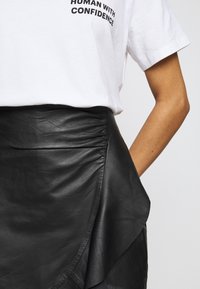 Zwarte leren rok met een wikkelontwerp en plooidetail bij de taille, gecombineerd met een witte katoenen t-shirt met vetgedrukte zwarte tekst.