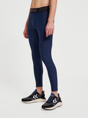 Marineblauwe sportlegging met een zwarte elastische tailleband waarop het "HUMMEL"-logo staat. Draagt zwarte en blauwe sportschoenen met witte accenten.