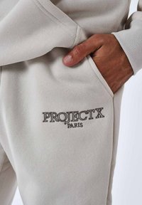 Pantalones deportivos de color gris claro con un acabado texturizado, que cuentan con un bolsillo lateral y un logo bordado "PROJECT X PARIS" en gris más oscuro.