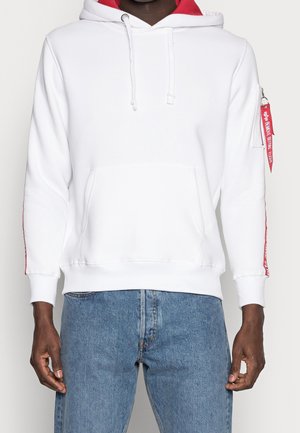 Mand iført hvid hættetrøje med rød inderside, rød tekst på ærmerne, frontlomme og blå jeans, stående mod en ensfarvet baggrund.