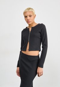 Svart ribbet to-delt antrekk med en cropped glidelåsjakke med lange ermer og et høyhentet blyantskjørt, strukturet stoff.