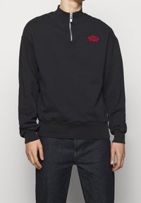 Black sweatshirt with high neckline and a quarter zipper. Features red embroidered logo on chest. Soft, textured fabric; ribbed cuffs and hem.