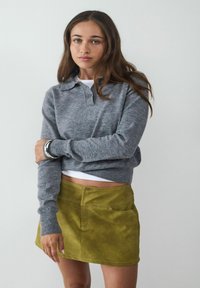 Grau gestrickter Pullover mit Kragen und Knopfverschluss, kombiniert mit einem leuchtend grünen Wildleder-Minirock, der über vordere Taschen und sichtbare Nähte verfügt.