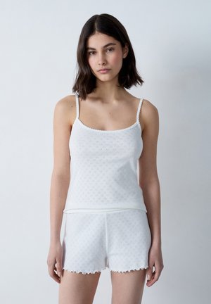 Camisola bianca in cotone con spalline sottili e design ad occhio di pernice, abbinata a pantaloni corti bianchi coordinati con bordo scallop e vita elasticizzata.
