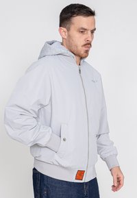 Hellgraue Bomberjacke mit Rei�ßverschluss, grauer Kapuze, elastischen Bündchen und einem orangenen Etikett am unteren Saum. Glatte Textur, lässiges Design.