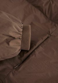 Bruine gewatteerde jas met een gathered mouwmanchet, geribbelde brei-detail en een zijrits. Glad, waterafstotend materiaal met een subtiele glans.