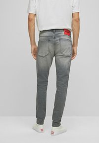 Jeans de mezclilla gris con corte slim, que cuentan con dos bolsillos traseros y una etiqueta roja con "HUGO" en la cinturilla. Llevados con zapatillas blancas.