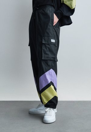 Person trägt schwarze Cargohosen mit lila und gelben Chevron-Mustern an den Unterschenkeln und weiße Turnschuhe, steht auf grauem Boden.