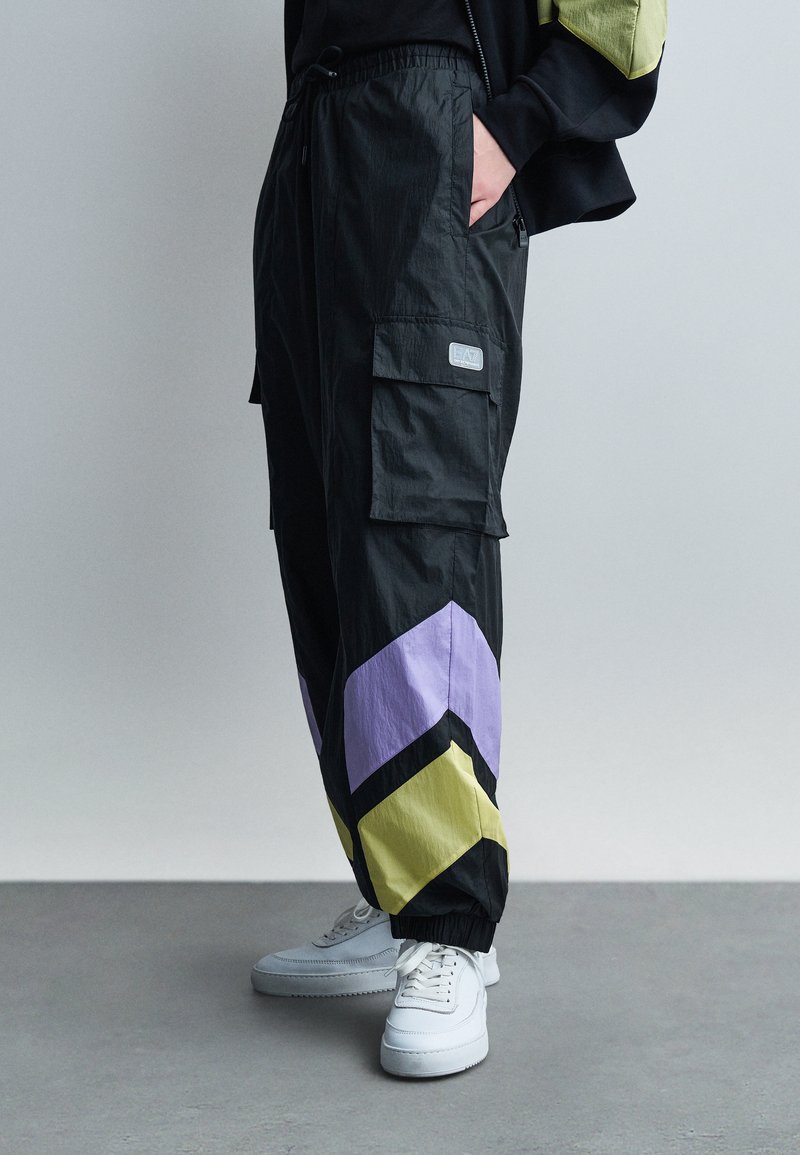 Άτομο που φοράει μαύρο παντελόνι cargo με μοτίβα βέλους σε μωβ και κίτρινο στο κάτω μέρος των ποδιών και λευκά αθλητικά παπούτσια, στέκεται σε γκρι πάτωμα.