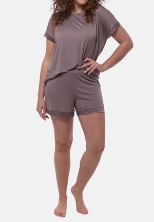 Kvinde stående barfodet iført en løs kortærmet top og matchende shorts i en afdæmpet mauve farve på en ensfarvet baggrund.