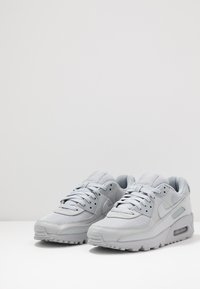 Pelēki sporta apavi ar sietu un ādas virsu, polsterētu apkakli un teksturētu zoli. Satur Nike zīmolu sānos un mēlē.