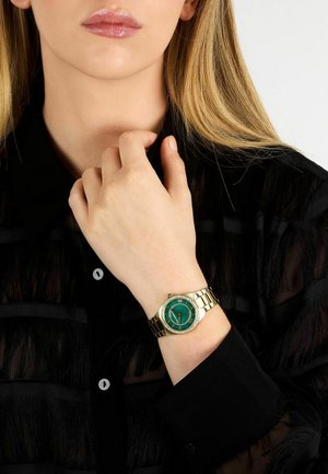 Guldarmbåndsur med rund grøn urskive, guldfarvet kædelink-armbånd og dekorative detaljer på rammen; båret på en hånd mod en sort skjorte.