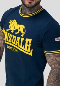 Muž v námořnickém modrém tričku Lonsdale London s žlutým logem lva a pruhovaným límcem a rukávy, ukazující tetované ruce a krk.