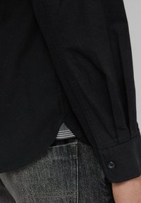 Schwarz geknöpftes Hemd mit glatter Textur, klassischem Kragen, langen Ärmeln und abgerundetem Saum; über grauen gemusterten Jeans getragen.