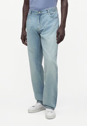 Mann trägt hellblaue, locker sitzende Jeans mit fünf Taschen und weißen Sneakers vor einfachem weißen Hintergrund.