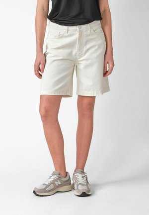Person iført off-white knælange shorts, sort top og grå New Balance sneakers, stående mod en ensfarvet lys baggrund.