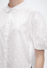 Bela srajca s kratkimi rokavi z teksturiranimi pikami, spredaj z gumbi, ohlapnimi rokavi in koničastim ovratnikom na osebi.