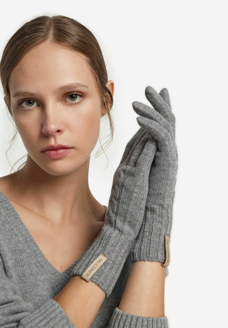 Gants tricotés gris en matériau doux avec des poignets côtelés et une étiquette au poignet. Les gants ont un design ajusté et épuré.