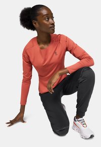 Haut de sport corail à manches longues avec un col rond, associé à un pantalon noir ajusté et des chaussures de course blanches avec des accents colorés.