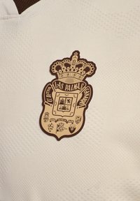 Weißer strukturierter Stoff mit einem braunen Wappenlogo mit Krone, das den Text "UD Las Palmas" und mehrere andere Schildsymbole zeigt.
