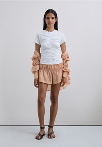 Mujer vestida con una blusa blanca corta con fruncidos, pantalones cortos de capas melocotón, una camisa suelta melocotón colgando de los brazos y sandalias negras con tachuelas.