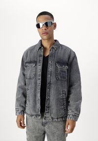 Chaqueta de mezclilla gris con dos bolsillos en el pecho, cierre de botones y costuras en contraste, combinada con pantalones de mezclilla gris y gafas de sol.