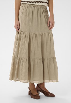 Žena oblečená v béžovej viacvrstvovej midi sukni, doplnenej hnedými tkanými sandálmi na podpätku so zapínaním za pätu, stojaca pred jednoduchým pozadím.