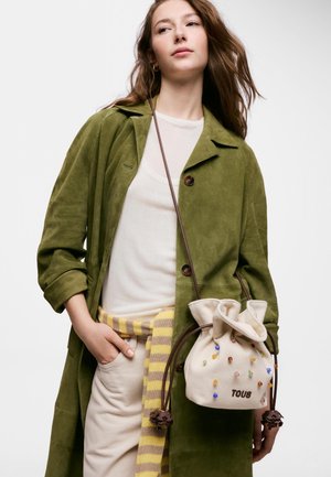 Femme portant un manteau vert olive et une tenue beige tient un sac beige à cordon avec des perles colorées et le logo "TOUS" sur le devant.