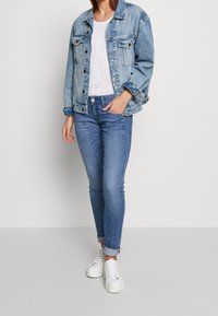 Světle modrá denimová bunda s dvěma náprsními kapsami, nošená přes bílý síťovaný top, spárovaná s úzkými modrými džíny a bílými teniskami.