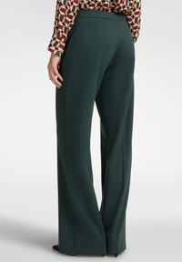Donna che indossa pantaloni larghi verde scuro e una blusa a fantasia geometrica colorata, in piedi con una mano rilassata al suo fianco.