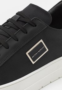 Adidași negri cu un superior texturat, șireturi plate și o talpă albă. Prezintă un accent logo metalic dreptunghiular pe lateral.