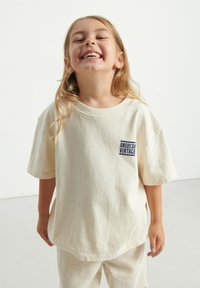 T-shirt en coton couleur crème à manches courtes, avec un logo bleu "American Vintage" sur la poitrine gauche. Texture lisse.