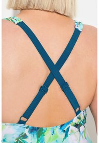 Les bretelles en X de couleur sarcelle se détachent sur un fond à motifs floraux. Les bretelles sont équipées de matériel ajustable pour un ajustement personnalisé.
